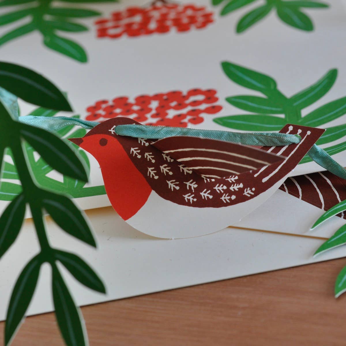 Close-up of a red-breasted paper robin threaded onto green ribbon beside rowan leaves and berries